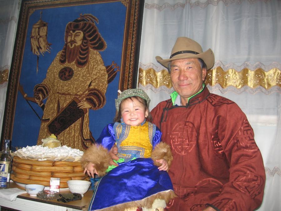An older Mongolian man in a cowboy hat and a rust-red silk deel smiles, holding a beaming toddler in a blue and gold deel on his lap, beside a table stacked with ul boov pastries, a woven wall hanging of a Mongol warrior behind them