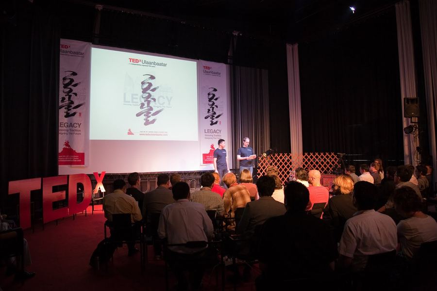 Two hosts in dark T-shirts, one holding a tablet, stand on stage before a seated audience at TEDxUlaanbaatar, beside giant red TEDx letters and under a screen showing the event’s Legacy theme