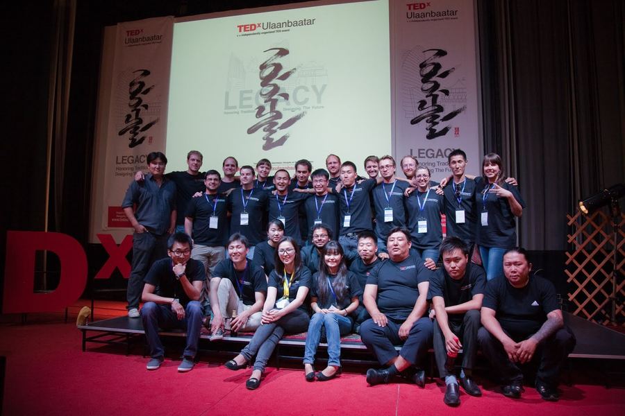 About thirty TEDxUlaanbaatar organizers and volunteers in matching black T-shirts and lanyards pose together on the stage in front of the Legacy screen and banners