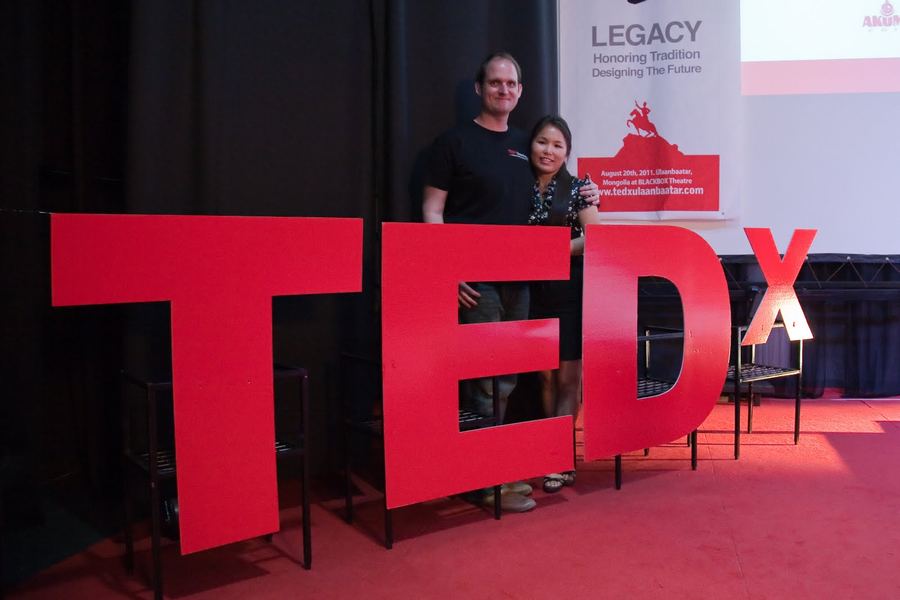 Travis and Tunga stand arm in arm behind giant red TEDx letters on the stage, next to a banner reading “August 20th, 2011, Ulaanbaatar, Mongolia at BLACKBOX Theatre”
