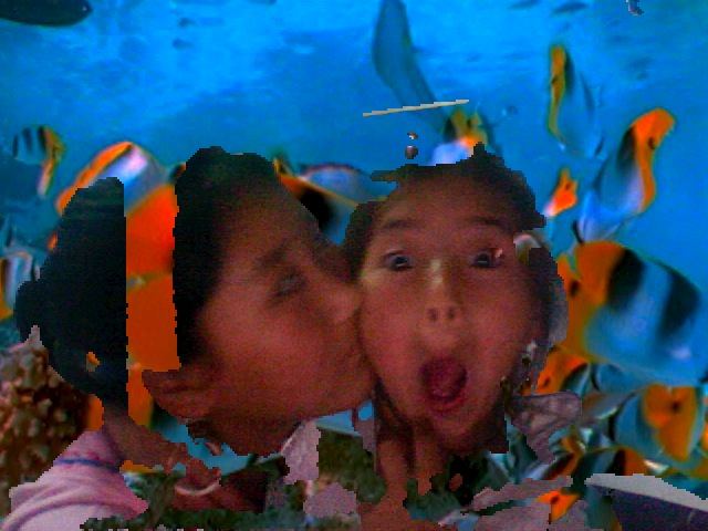 An older girl kissing the cheek of a young girl who makes a surprised face, in a Photo Booth picture with a backdrop of tropical fish