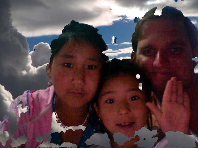 Travis waving beside an older girl and a young girl, posing cheek to cheek in a Photo Booth picture with a cloudy sky backdrop