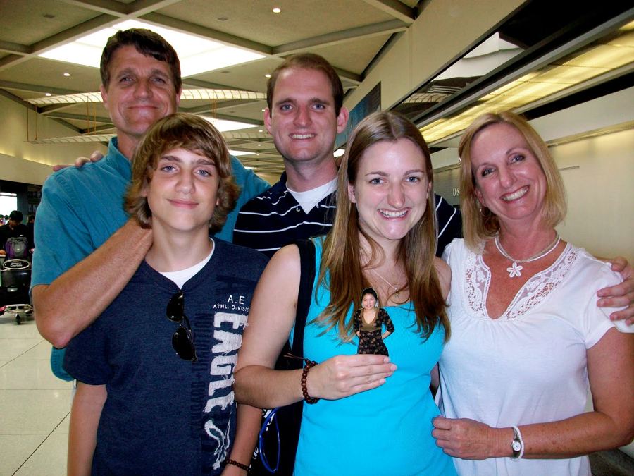 Travis and four family members smile together in an airport arrivals hall; the young woman in front holds up a small cutout photo of Tunga