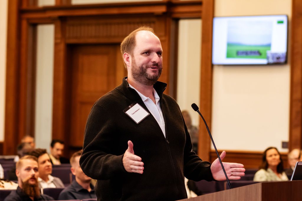 Travis Hellstrom, in a black sweater and name tag, smiling as he speaks at the podium in the Virginia General Assembly chamber